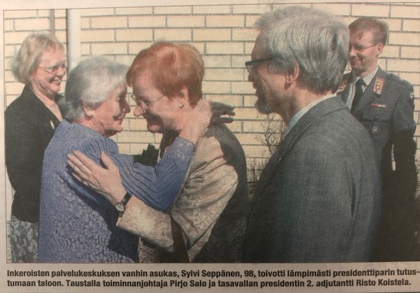 Presidentti Tarja Halonen puolisonsa kanssa vierailulla, hän halaa ulkona iäkästä hymyilevää rouvaa. Taustalla Kymijoen Hoivan toiminnanjohtaja sekä presidenttiparin adjutantti. 